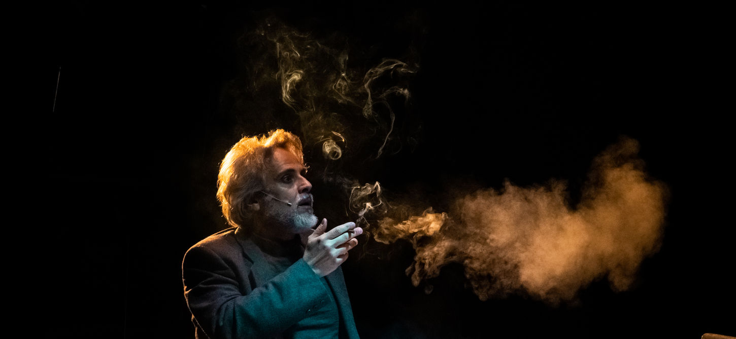 Nella fotografia c'è un uomo seduto, ha un tavolo davanti e sta fumando una sigaretta e c'è un po' di fumo. Lui è leggeremente illuminato e lo sfondo è nero. Lo spettacolo andrà in scena al Teatro Bobbio de La Contrada Teatro Stabile di Trieste.