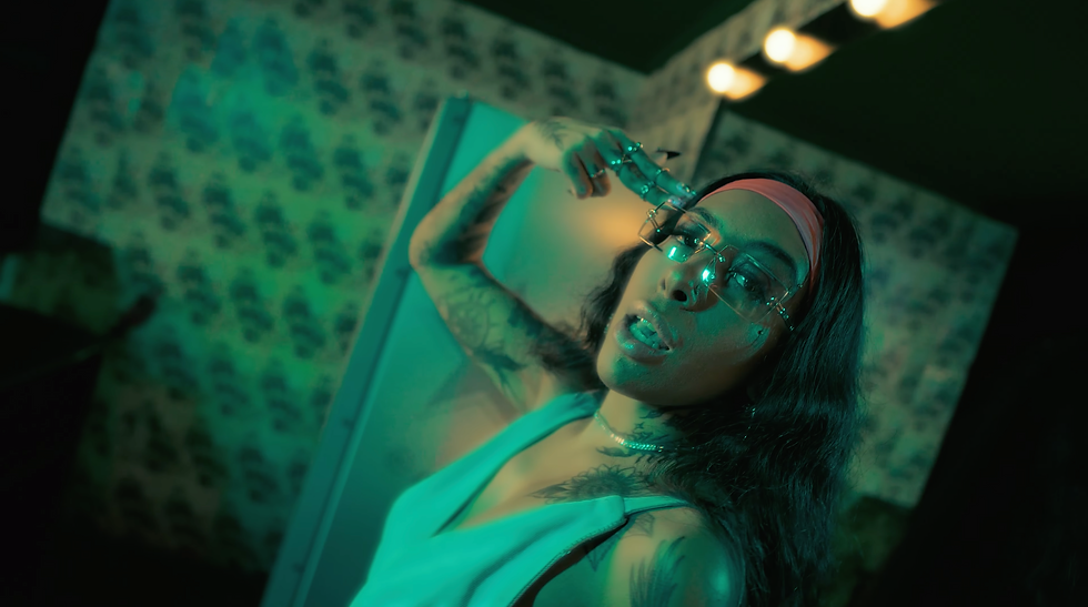 Mari raia está cantando, ela usa um oculos de grau e uma faixa rosa no cabelo, está cantando para seu videoclipe "Até o Amanhecer"