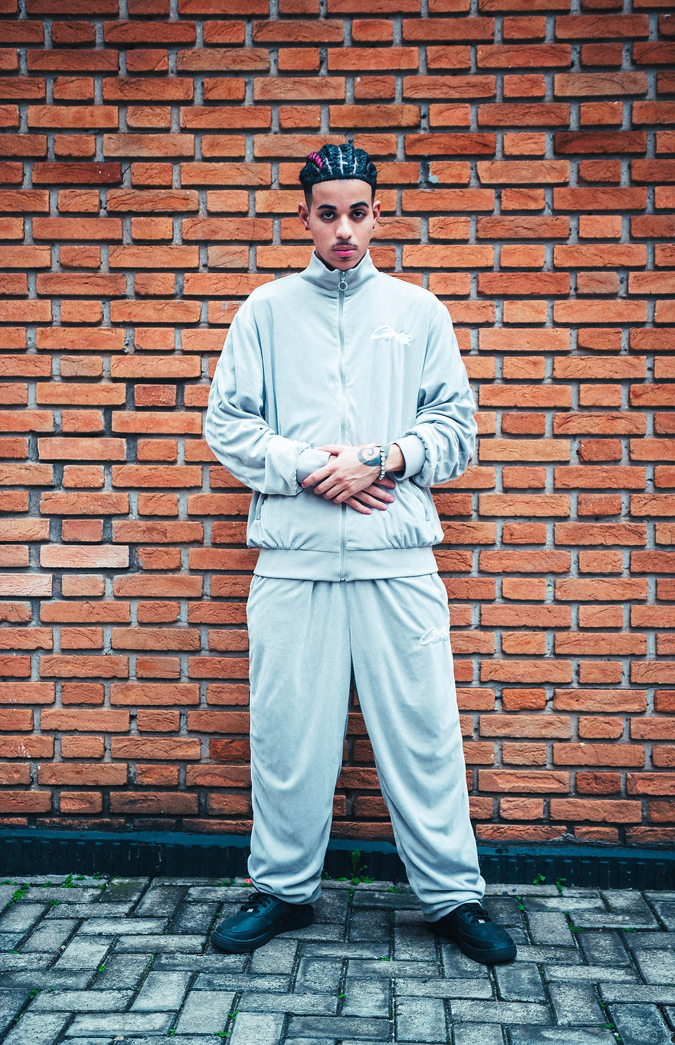 Lago veste um conjunto esportivo, casaco e calças cinza claro. Ele está em frente a um muro de tijolos, na rua