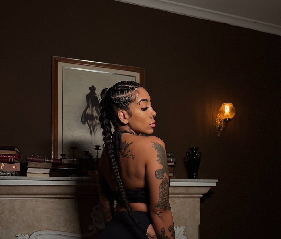 Mari Raia veste um vestido preto e usa tranças, ela está em frente a uma lareira com decorações em cima. O ambiente é escuro em tons terrosos.