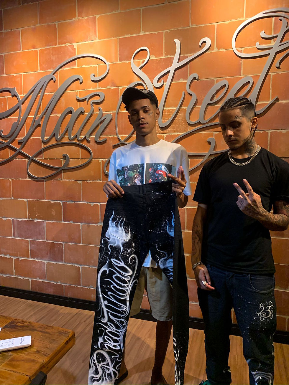Zick está vestindo sua marca de roupas, ao lado está o artista Bin segurando uma de suas peças exclusivas. Ambos estão em frente ao letreiro da Mainstreet gravadora do Rio de Janeiro