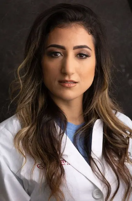 Woman in a white lab coat with long hair and a neutral expression against a dark background. Coat features faint red text.
