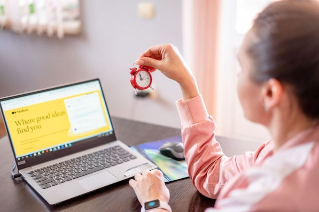 A woman sitting at desk with a laptop computer holding a small clock
