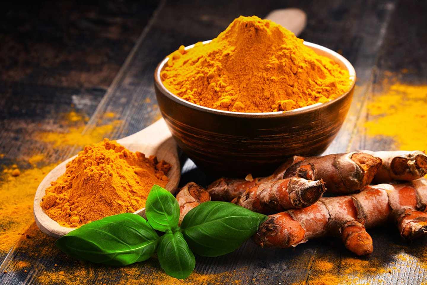Bowl and spoon with turmeric powder on dark wood. Fresh turmeric roots and green leaves nearby, with vibrant orange and green hues.