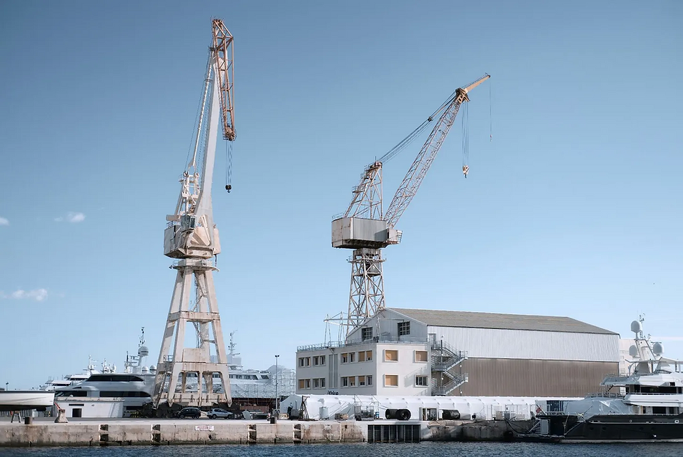Two tall harbor cranes beside yachts and a warehouse in a shipyard under a clear blue sky.