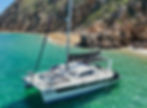 Catamaran "Love Life" on turquoise water near rocky coast and sandy beach with people. Clear sunny day, relaxed mood.