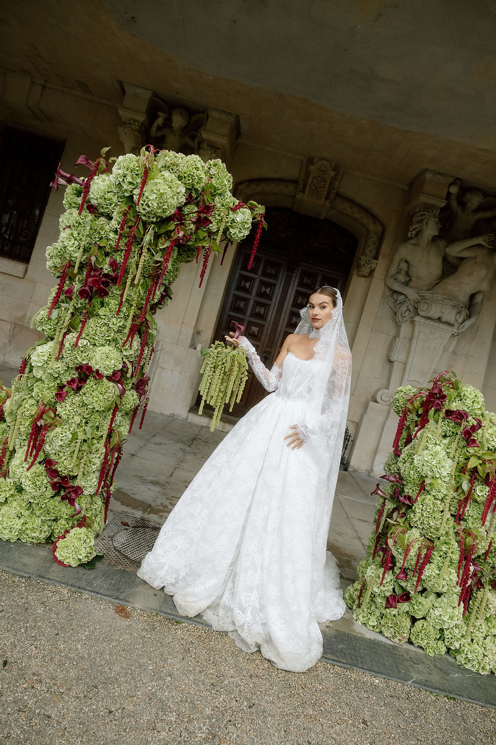 Mariée posant contre une structure florale d'hortensias verts et callas bordeaux, entrée sculptée du Château Bouffémont