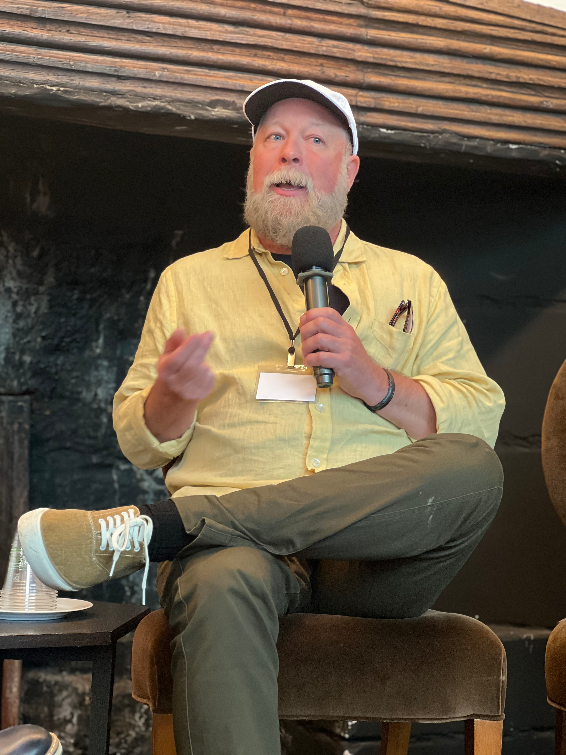 A panelist in a yellow shirt and white cap speaking into a microphone while seated, gesturing with one hand during the panel discussion.