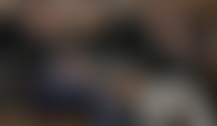 Two men wearing THE REAL branded caps sit on a stage in front of a fireplace, both holding microphones and pointing toward the audience during an energetic fireside chat conversation.