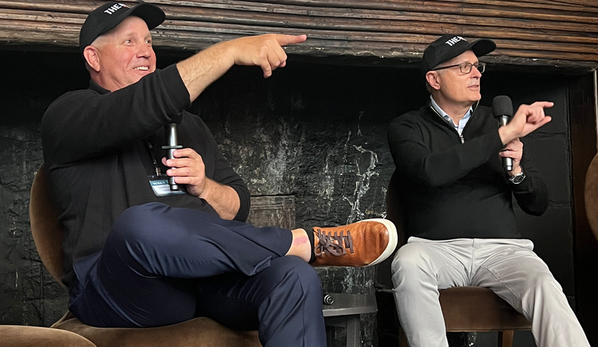Two men wearing THE REAL branded caps sit on a stage in front of a fireplace, both holding microphones and pointing toward the audience during an energetic fireside chat conversation.