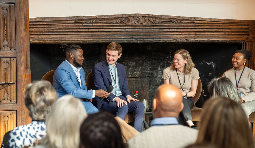Four panelists engaged in a panel conversation while seated by a large stone fireplace.