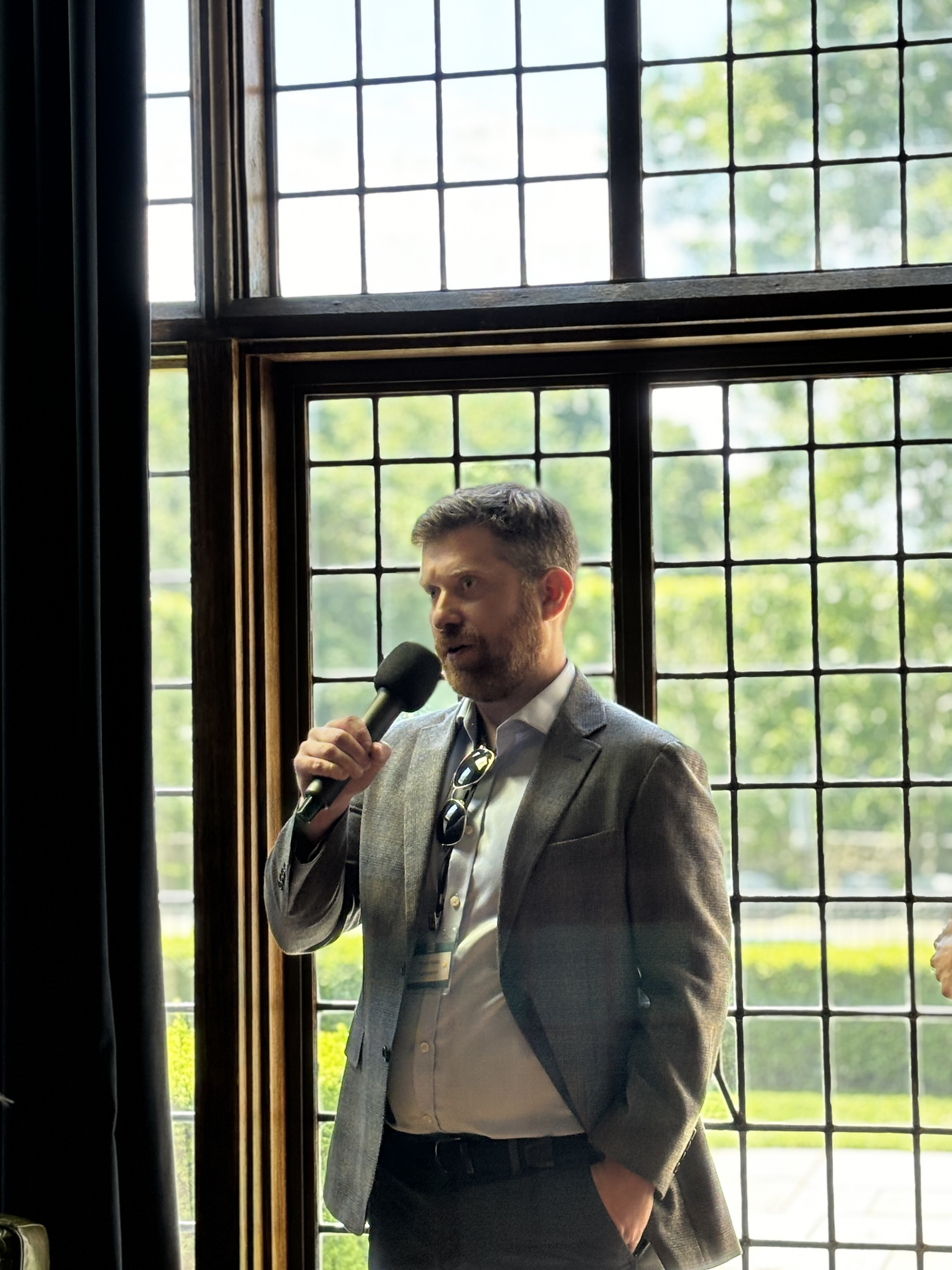 An attendee standing in front of large leaded glass windows, holding a microphone and addressing the room with a question for the speakers.