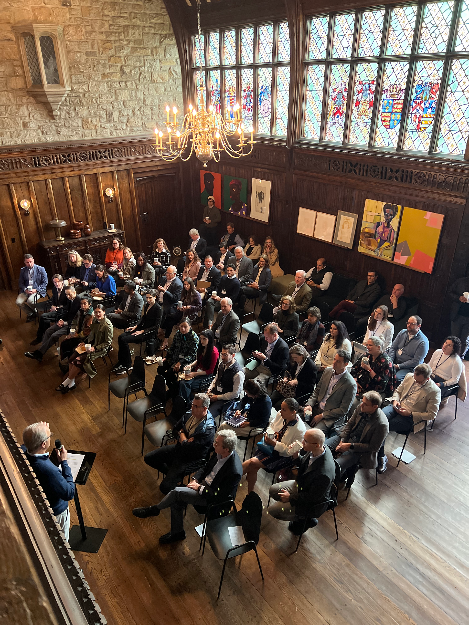 An overhead view of a large room with a high ceiling, showcasing an audience seated in rows facing a speaker on a small stage to the left. The room features intricate dark wood paneling, large windows with stained glass designs depicting crests, and a large chandelier hanging from the ceiling. Artwork is visible on the far wall.