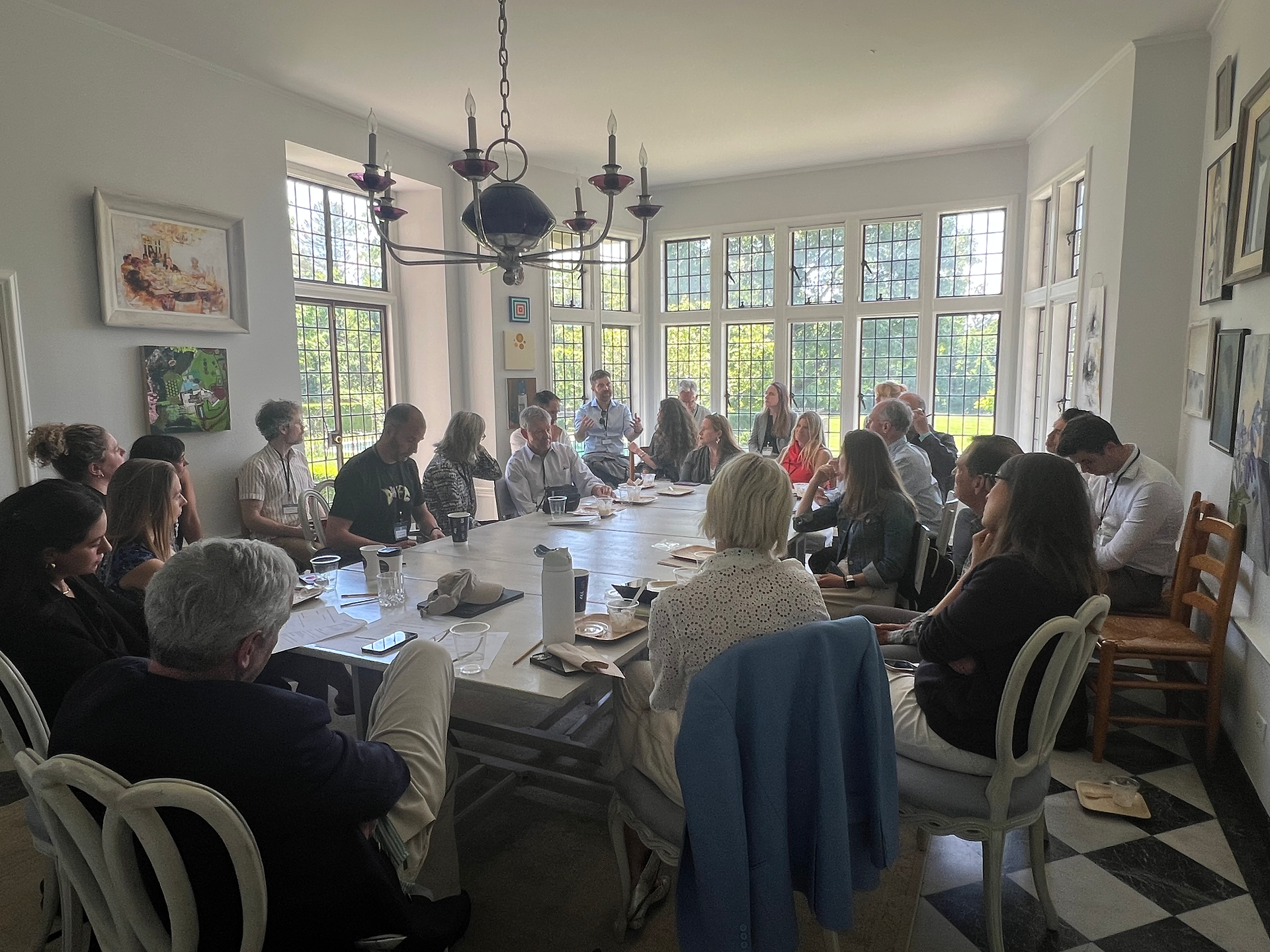 A large group of attendees seated around a long table in a bright room with leaded glass windows, engaged in a breakout working session.