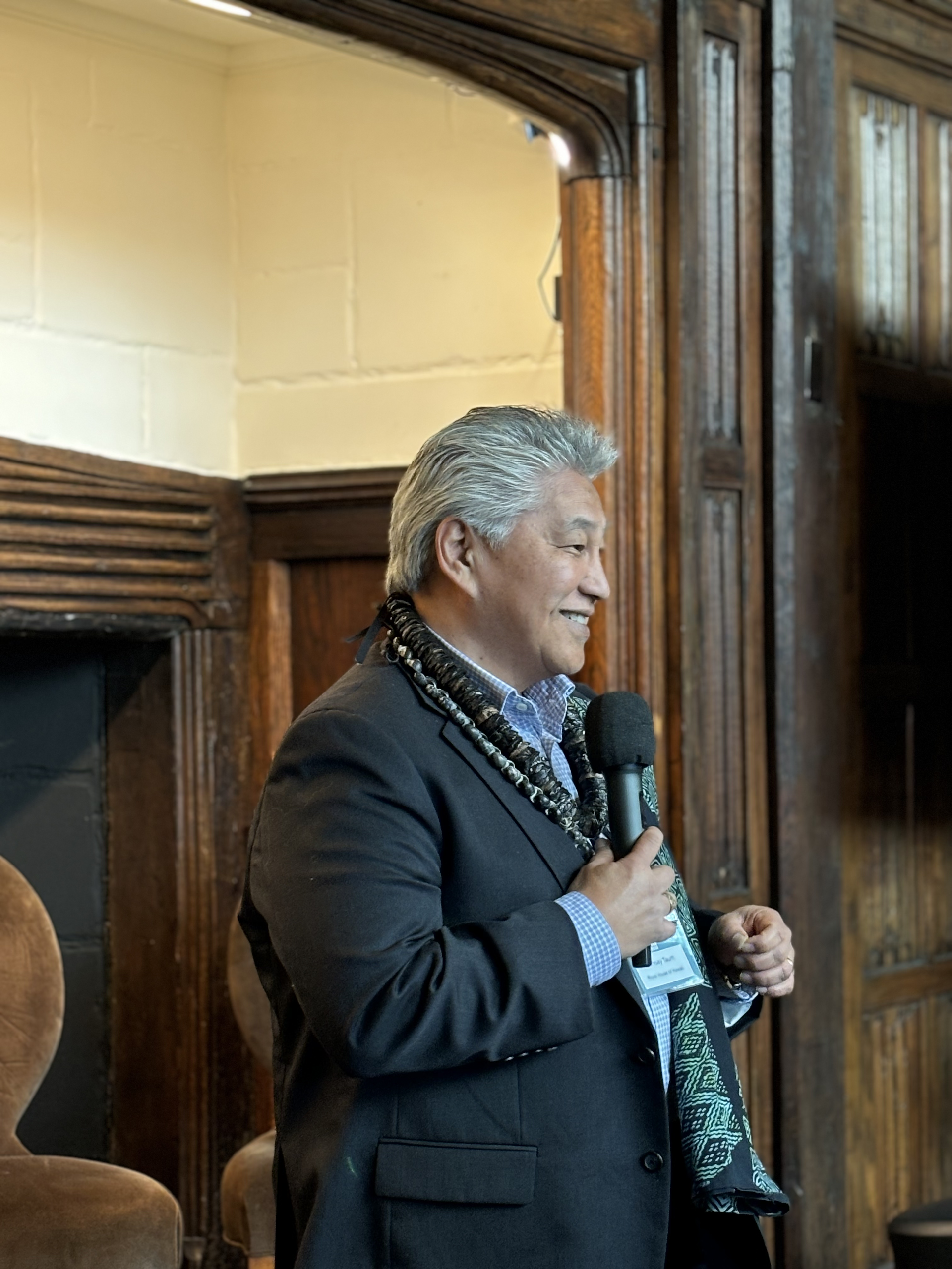 A male speaker in a dark blazer and traditional beaded necklace holds a microphone while speaking at Foundation House, with wooden architectural details visible in the background.