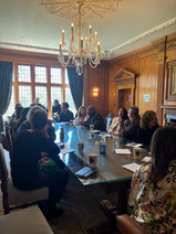 Attendees gathered around a polished conference table in an elegant room with wooden paneling, crystal chandelier, and large windows overlooking the grounds.