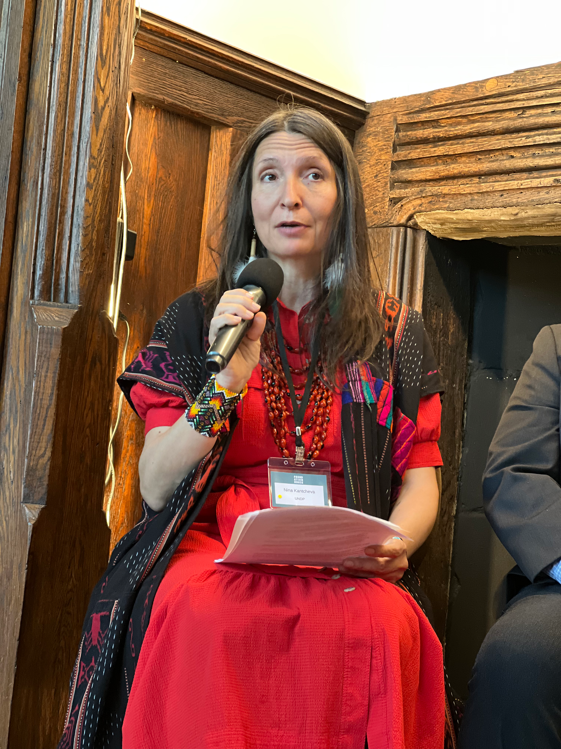 A female speaker wearing traditional Indigenous textiles in red and black, with beaded jewelry, holds a microphone and notes while speaking in front of wooden architectural details.