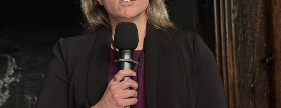 A woman with short blonde hair, wearing a black blazer and a lanyard, stands holding a microphone and papers while speaking at an event. The background features a dark ornate frame or wall.