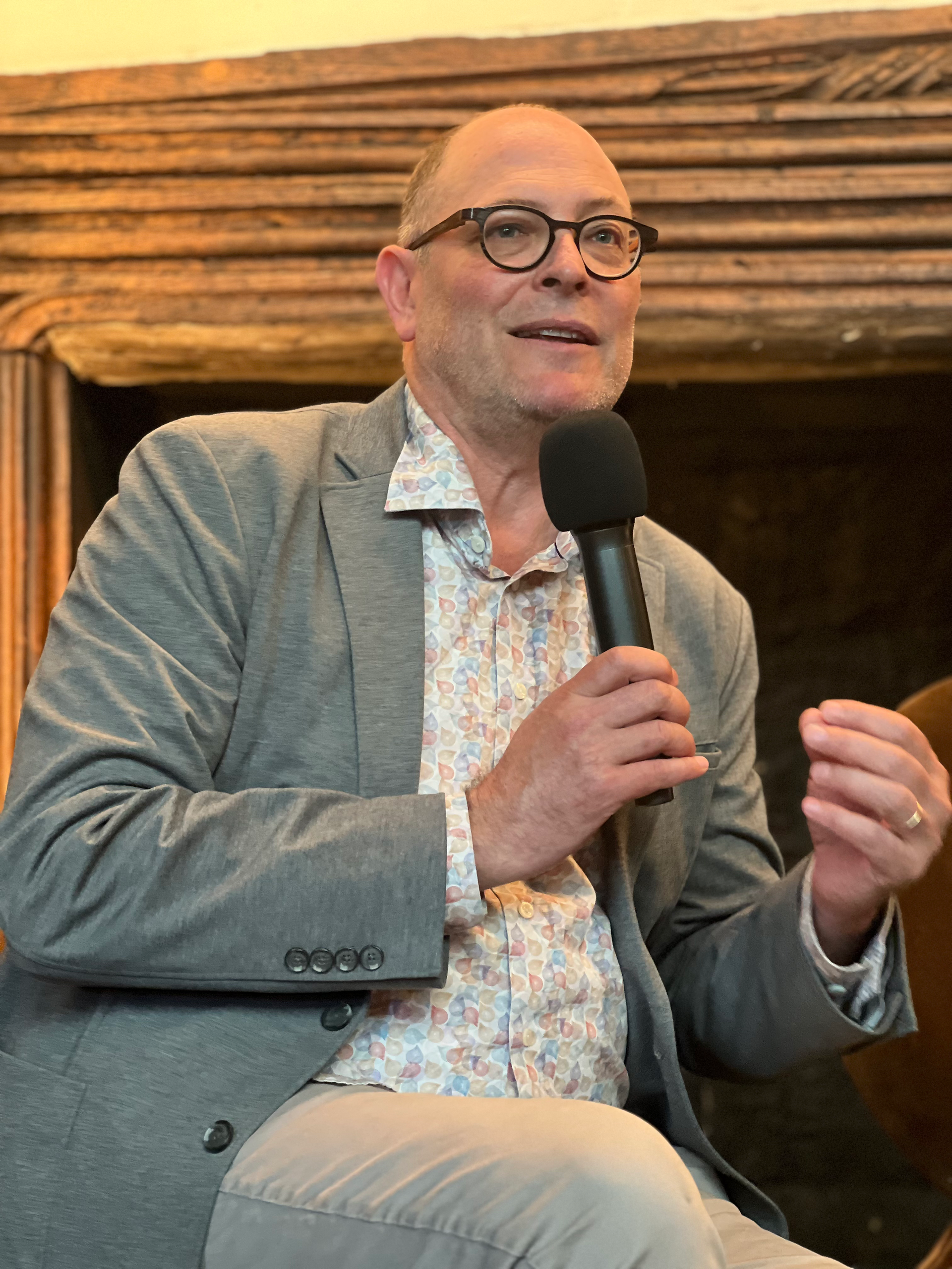 A panelist speaking into a microphone while seated, wearing a patterned shirt and grey blazer, engaged in conversation during the panel.
