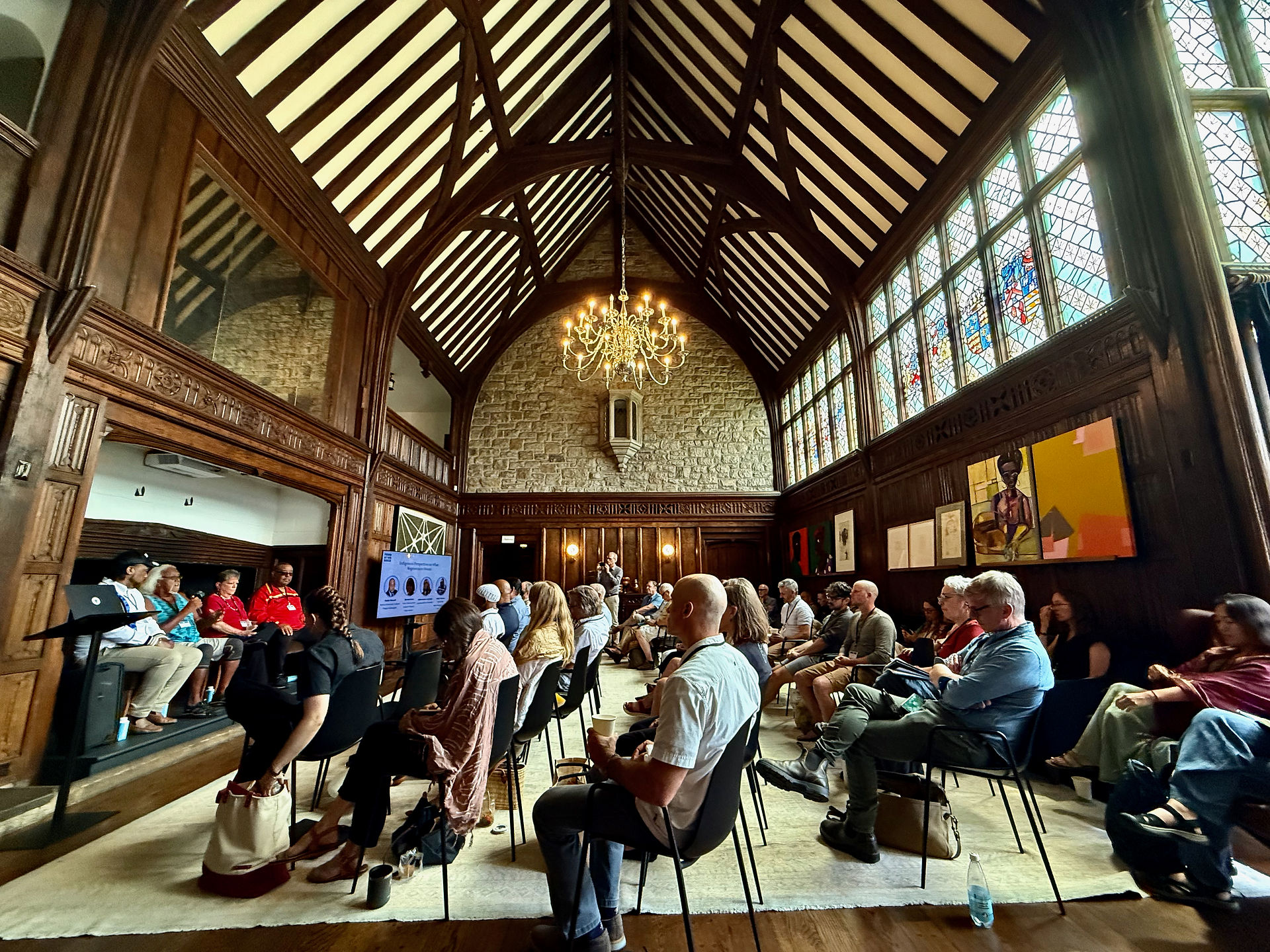A wide-angle interior shot of Foundation House's Great Room with soaring wood-beamed ceilings, stained glass windows, and a chandelier. A large audience sits in rows of chairs facing a presenter and screen at the far end of the room.