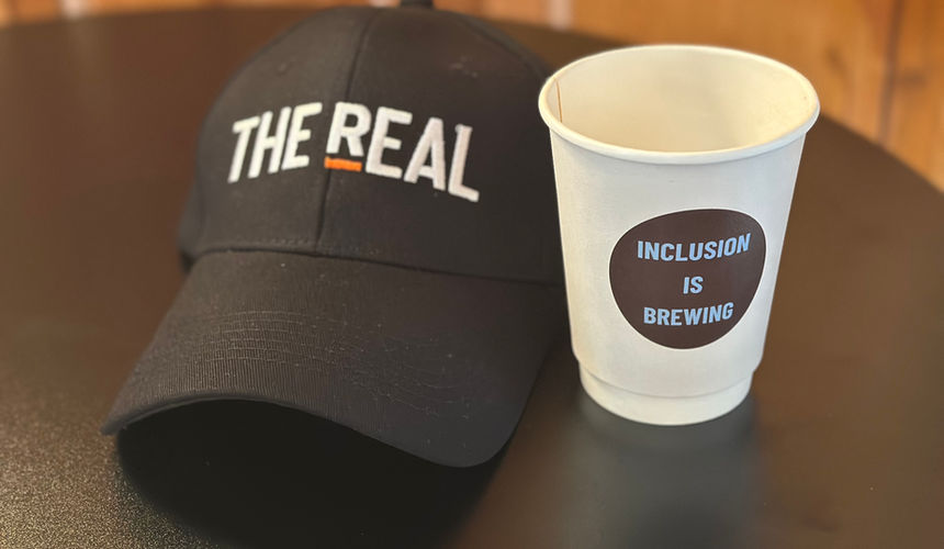 A black baseball cap embroidered with THE REAL logo in white and orange sits on a dark table next to a white paper coffee cup bearing a dark circular sticker that reads 'Inclusion is Brewing' in blue text.