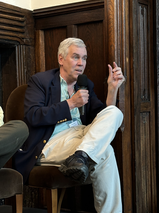 A panelist speaking into a microphone while seated on stage, gesturing with one hand during the panel discussion.