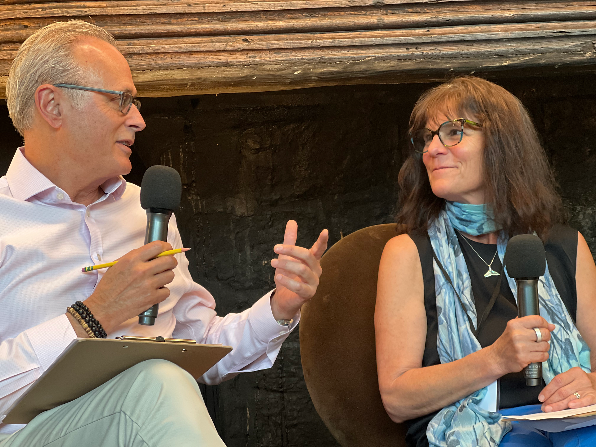 Two speakers in conversation on stage, one holding a clipboard and pencil, the other gesturing while speaking into a microphone during a fireside chat.
