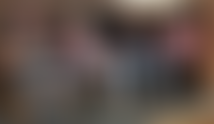 Four THE REAL Ambassadors wearing THE REAL branded caps sit in chairs on a raised platform in front of a ornate fireplace, each holding a microphone. The man on the left speaks animatedly while gesturing with his hand during the panel discussion.