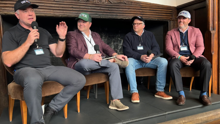 Four THE REAL Ambassadors wearing THE REAL branded caps sit in chairs on a raised platform in front of a ornate fireplace, each holding a microphone. The man on the left speaks animatedly while gesturing with his hand during the panel discussion.