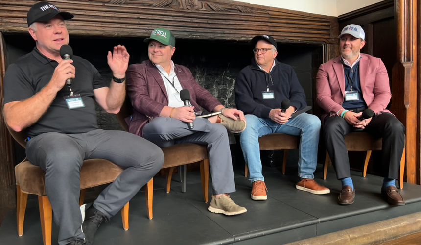 Four THE REAL Ambassadors wearing THE REAL branded caps sit in chairs on a raised platform in front of a ornate fireplace, each holding a microphone. The man on the left speaks animatedly while gesturing with his hand during the panel discussion.