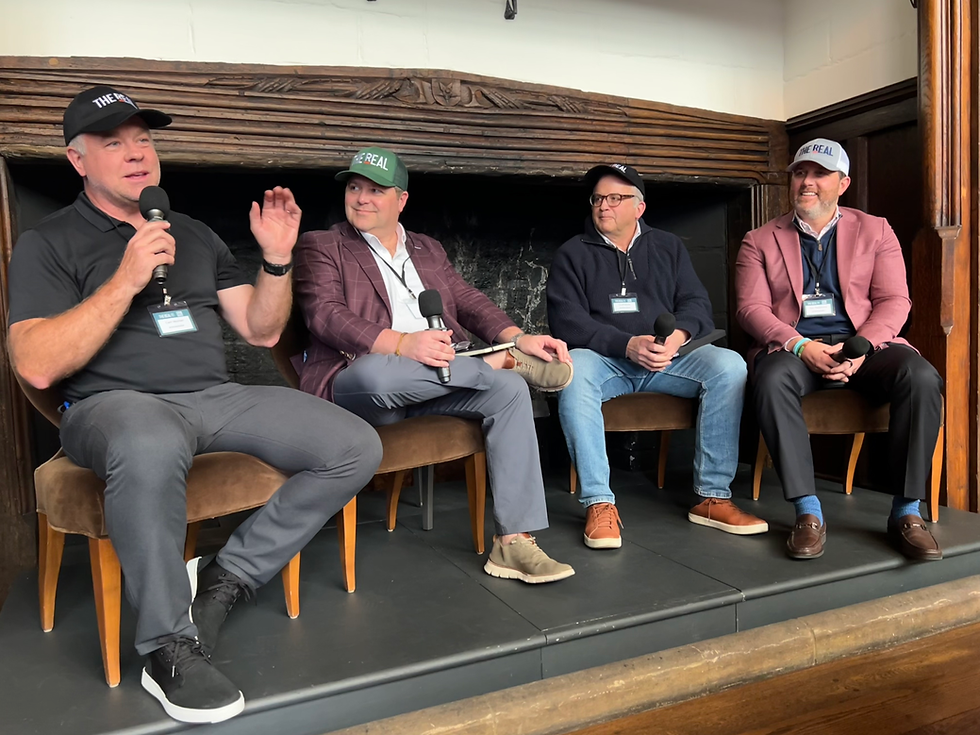 Four THE REAL Ambassadors wearing THE REAL branded caps sit in chairs on a raised platform in front of a ornate fireplace, each holding a microphone. The man on the left speaks animatedly while gesturing with his hand during the panel discussion.