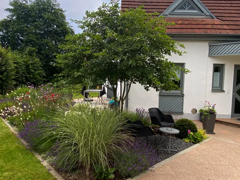 Gemütlicher Sitzplatz für den ersten Kaffeee in der Morgensonne im Vorgarten unter einem mehrstämmigen Baum (Pflaumendorn) und umgeben von bunt blühenden Stauden und Gräsern