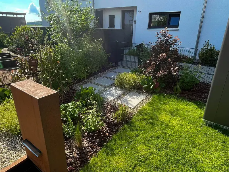Brunnen aus Cortenstahl und von Beeten umgebene Gartentür und Trittplatten