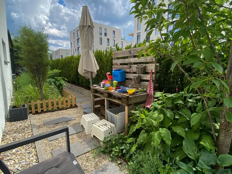 von Pflanzen umgebene Matschküche für Kinder, Sitzplatz an der Hauswand und Kiesweg mit Trittplatten
