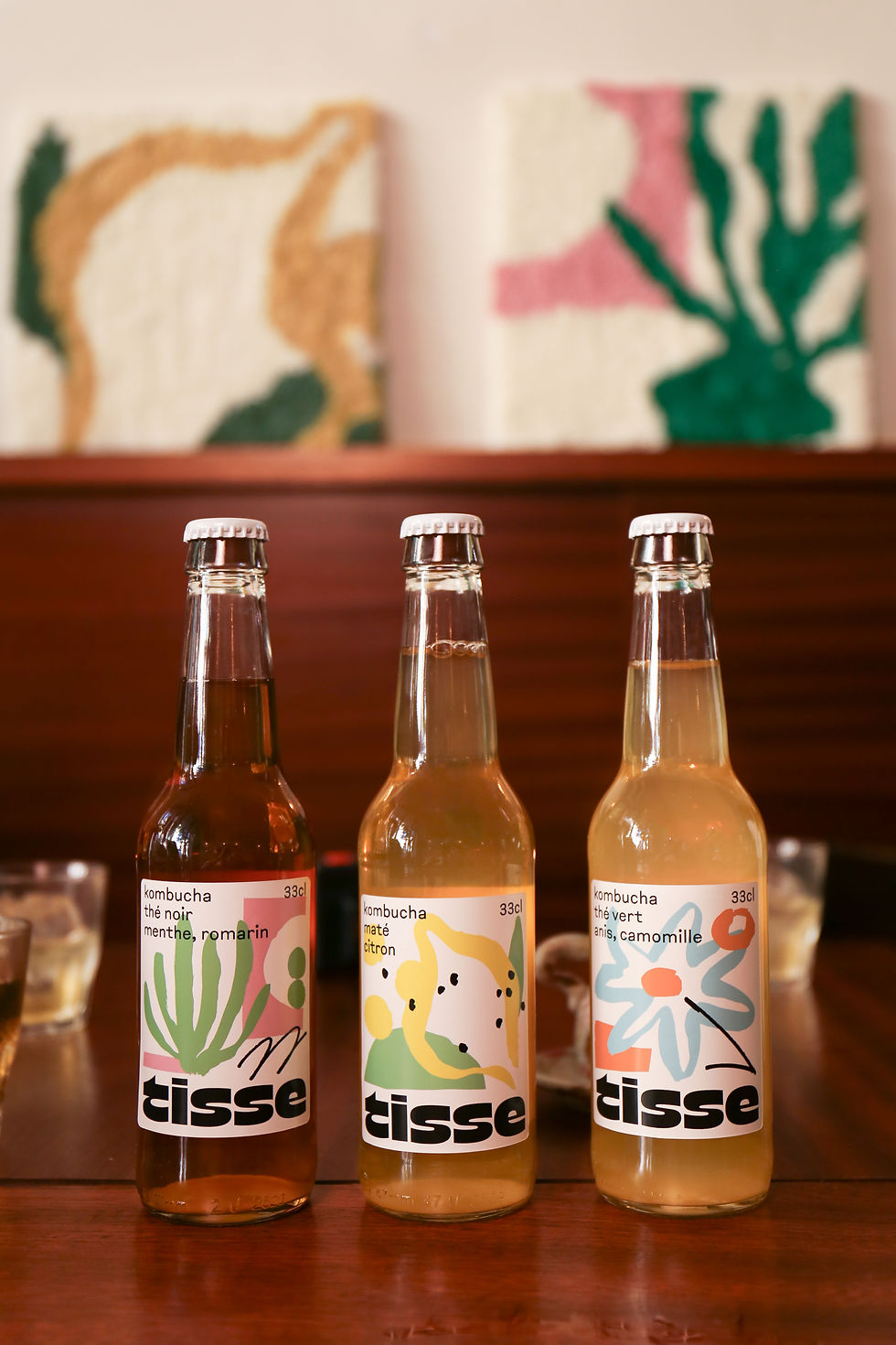 Trois bouteilles de kombucha Tisse aux saveurs thé noir menthe romarin, maté citron et thé vert anis camomille, posées sur une table en bois avec des verres en arrière-plan. L'étiquette colorée et artistique met en avant l'identité visuelle de la marque.