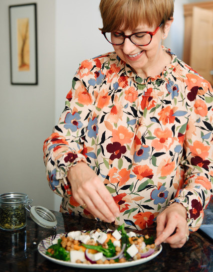 Smiling woman in red glasses and floral blouse plates a colorful salad in a bright kitchen.