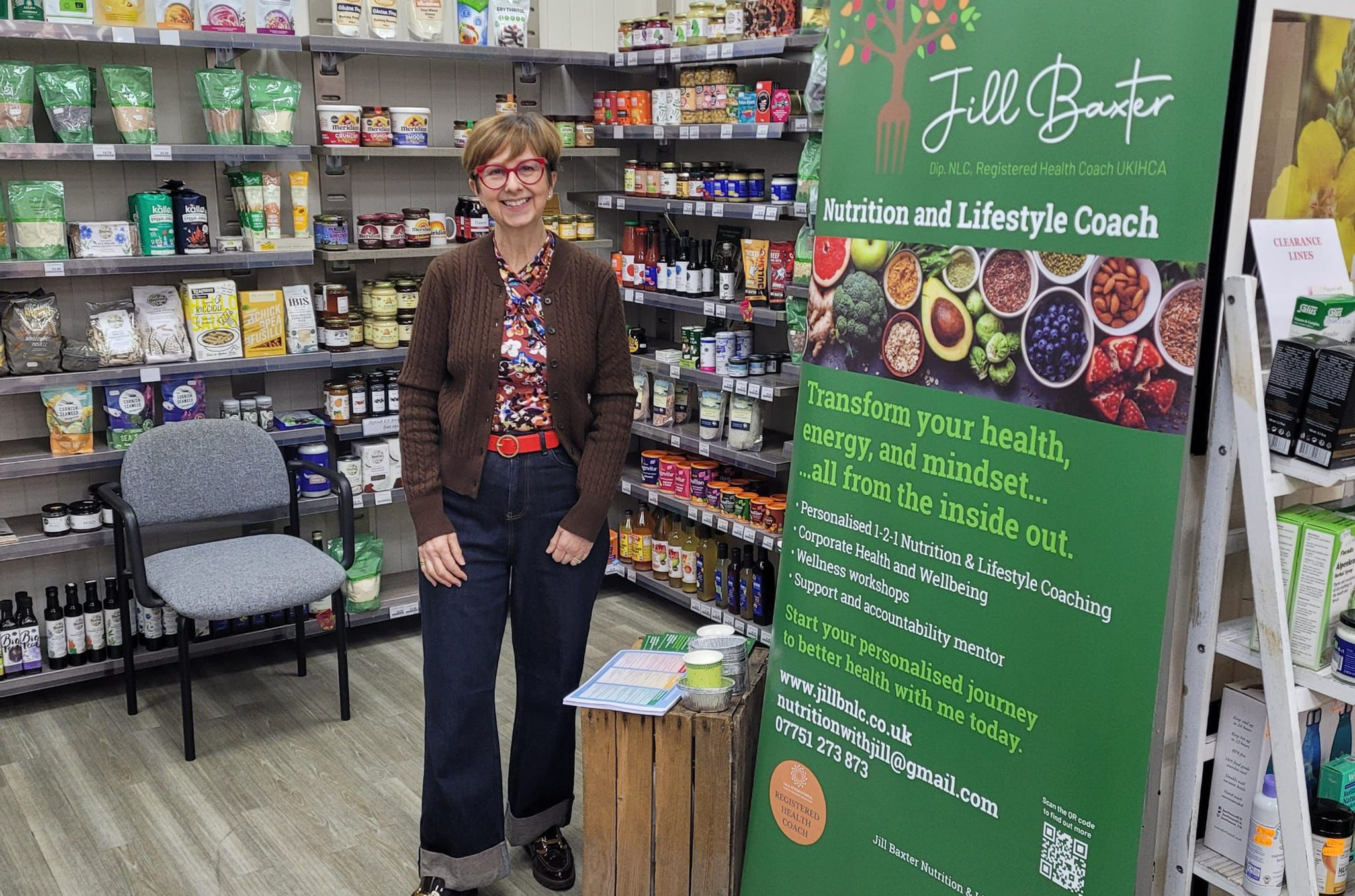 Smiling woman stands in a health food shop beside a green Jill Baxter Nutrition and Lifestyle Coach banner.