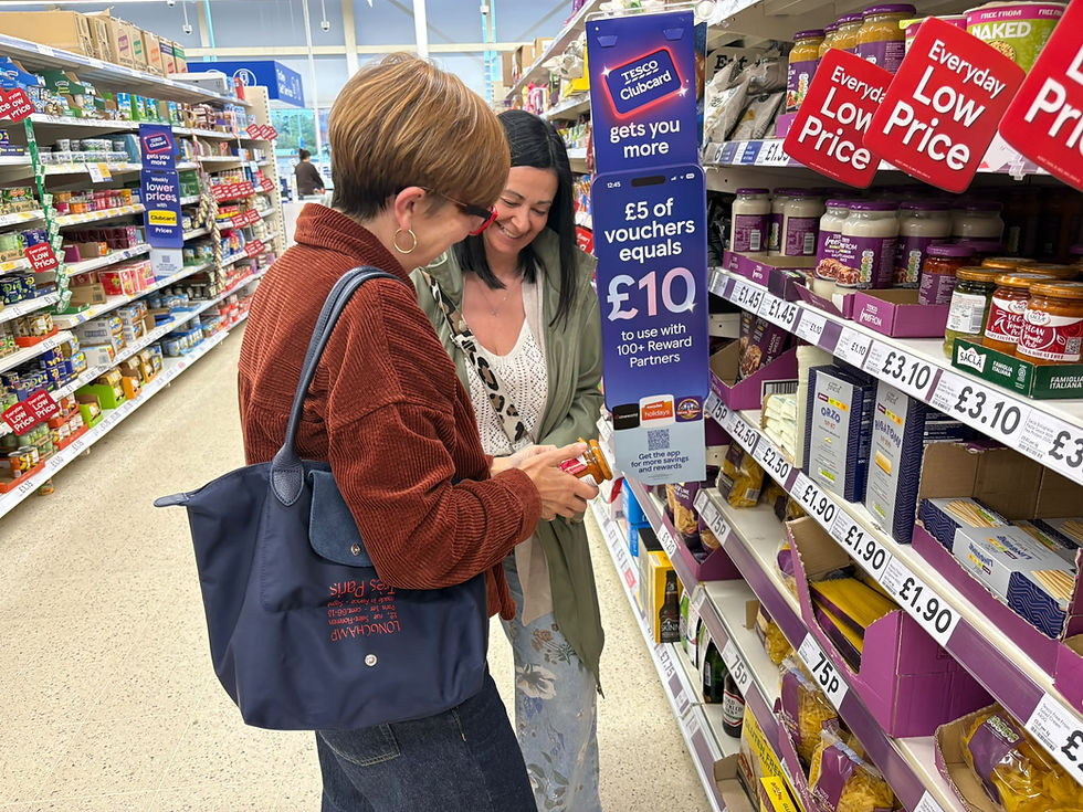 Two women smile in a Tesco grocery aisle, checking a phone and vouchers beside low-price signs and jars.