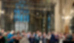 A picture of a choir consist mainly of people aged 50 plus, at the front of a traditional church which a large stain glass window behind the choir.