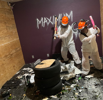 Two rage room session participants posing with bats and safety gear with items they smashed all around at the Maximum Rage LLC rage room in Humble near Houston, TX