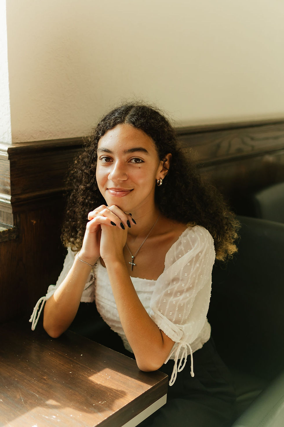 Yari smiling warmly at the camera, sitting at a table in a cozy coffee shop with hands resting under the chin