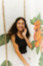 Yari sitting casually against a wall, smiling warmly at the camera in a professional portrait.