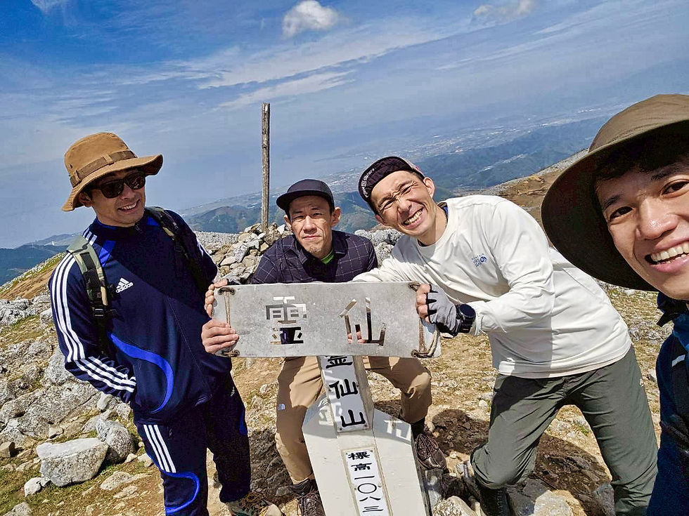山頂で4人が看板を持って笑顔。晴れた空と山並みを背景に、リラックスした雰囲気。看板には「雷」と書かれている。