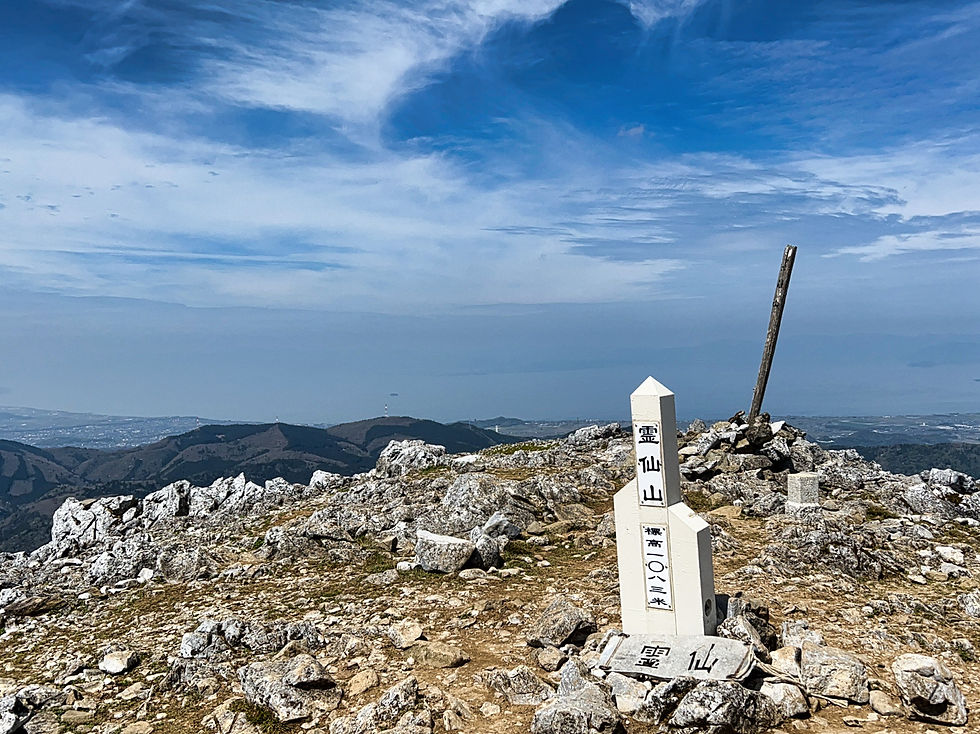青空の下、岩が散らばる山頂に「標高1,037メートル」と書かれた白い石碑が立つ。遠景には山並みが広がる。