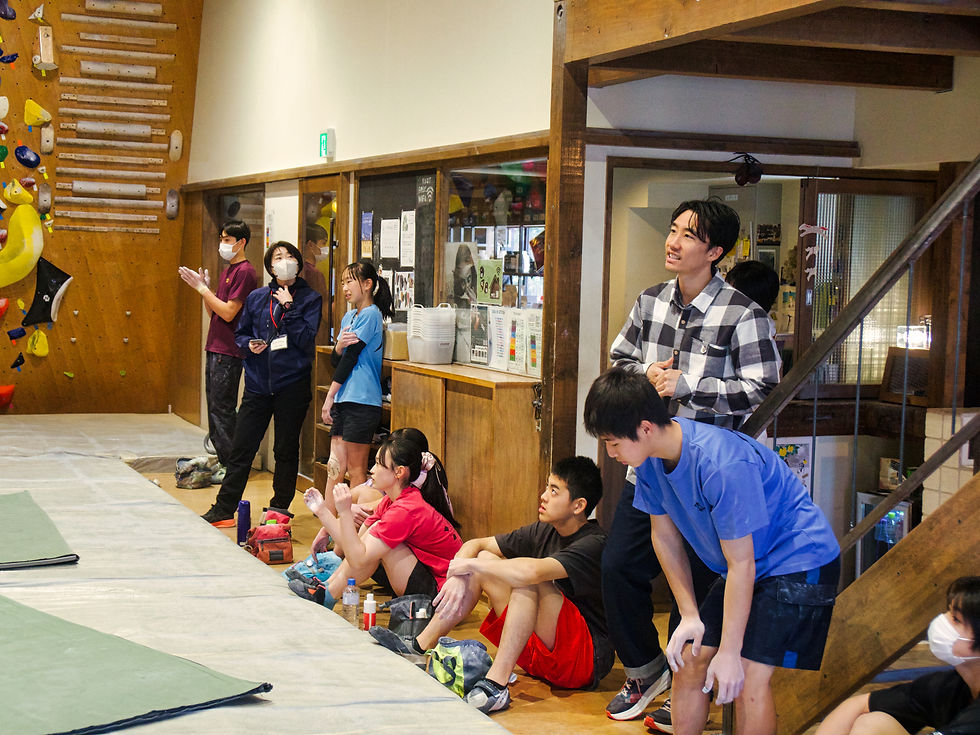 ボルダリングジムで複数の人々が壁を見ながら応援している。床に座った人と立っている人がおり、カラフルなクライミングホールドが背景に見える。