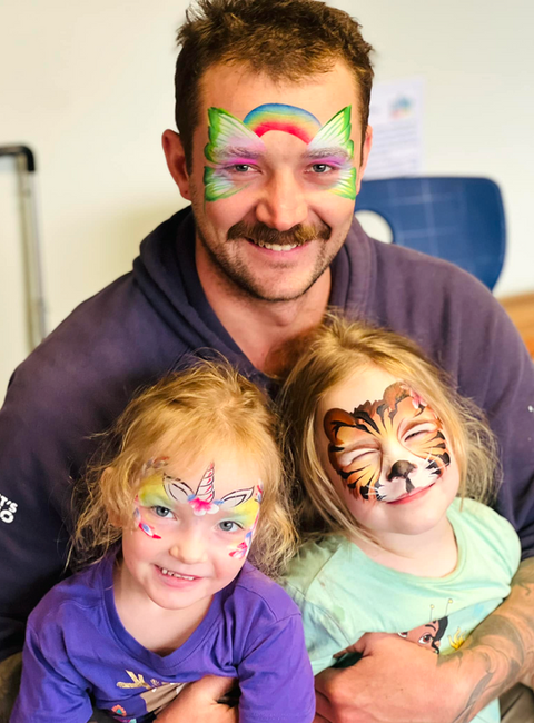 A smiling man and two girls with colourful face paint in the shape of different animals including unicorn, tiger and butterfly