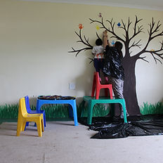 Volunteers painting a classroom wall while children imprint their palms with paint.