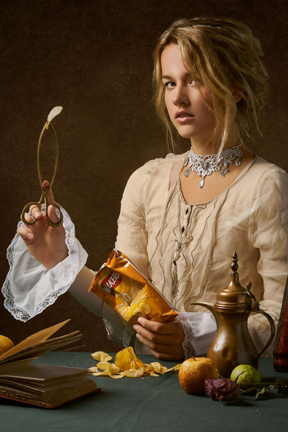 A woman in vintage attire holds a crisp with scissors. Chips and a book are on a green table, with fruit and a bronze pot. Moody setting.