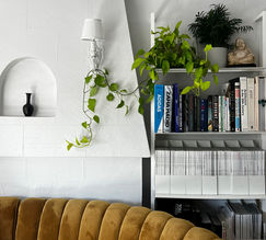 Living room vignette featuring a rounded, yellow, velvet sofa and the Temple Street Bookshelf by Pottery Barn in white, displaying a collection of indoor plants, coffee table books and various issues of Architectural Digest magazine. Professionally Organized and curated by The Neat Freak L.A. 