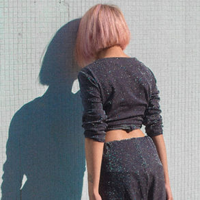 Femme aux cheveux rose pâle, dos tourné, en tenue sombre scintillante, devant un mur carrelé blanc avec grande ombre.